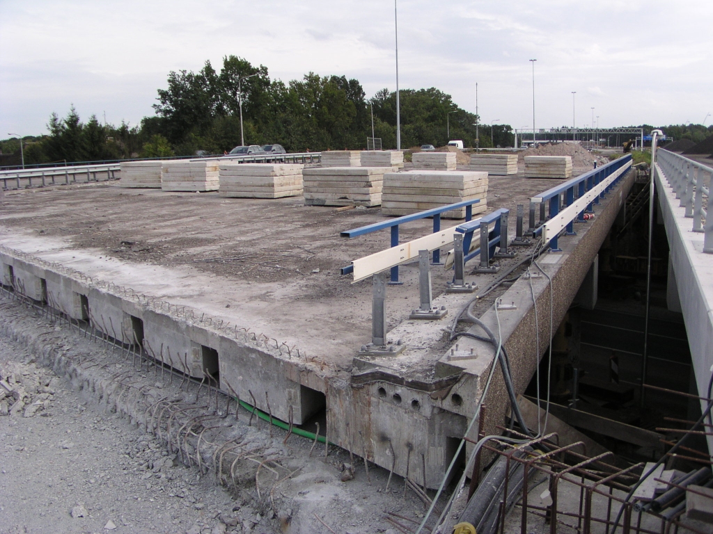 p9020004.jpg - Het sinds enige weken vacante viaduct in de westelijke oude hoofdrijbaan over de Oirschotsedijk is gestript. Tot onze vreugde zien we dat ook de blauwwitte brugleuning wordt verwijderd en hopelijk wordt vervangen door dezelfde strakke exemplaren als op de nieuwe viaducten (rechts). Te vrezen valt wel dat de grindtegel zijbeplating blijft zitten. Goed te zien de prefab ligger constructie met relatief dunne opvullaagjes ertussen. Het moet een huzarenstukje worden deze kolos op te vijzelen zonder dat er breuken ontstaan in die dunne gedeelten.