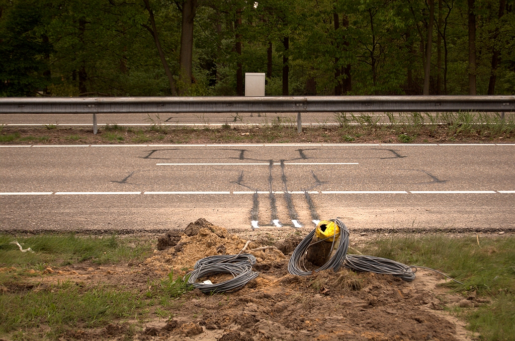 20090425-171143.jpg - Het zijn de detectielussen, een essentieel onderdeel van de signaleringsinstallatie. In de vernieuwde Randweg Eindhoven worden deze lussen in dicht asfalt gefreesd, en daarna door de ZOAB deklagen aan het zicht onttrokken. Men zal in bestaande wegvakken niet een complete ZOAB vervanging uitvoeren voor het wegwerken van een paar detectielusjes, met deze wonden in het wegdek als resultaat. De zwarte buis die uit de grond steekt zal dienen voor het leggen van de kabels van detectielussen naar het kastje aan de overzijde van de rijbaan, via een geboorde tunnel. Ook de witte kasten bij de portalen bevinden zich alle aan de westzijde van de rijbaan, zodat het doorgaande transportnetwerk van de signaleringsinstallatie zich daar bevindt. Kennelijk is de oplossing met geboorde tunnels praktischer dan het aanleggen van netwerk en kasten aan beide zijden van de rijbanen.