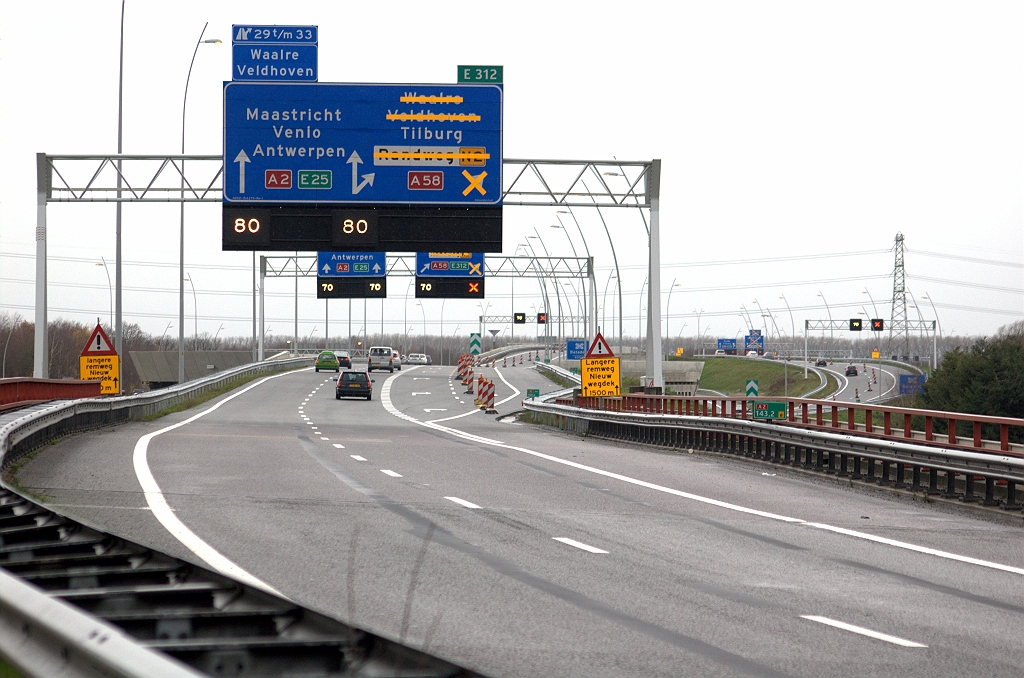 20091206-141422.bmp - Wel flinke wijzigingen aan het eerste beslisbord, zodat het nu duidelijk is dat er moet worden gekozen tussen de A58 en de A2, voorlopig inclusief de lokale doelen, waarvoor men een nieuw ruitertje heeft laten maken. Daarbovenop het afritnummerbordje dat tijdelijk is verplaatst van rechts naar links. E-nummer op het rechter ruitertje helaas nog steeds in het verkeerde lettertype.  week 200911 