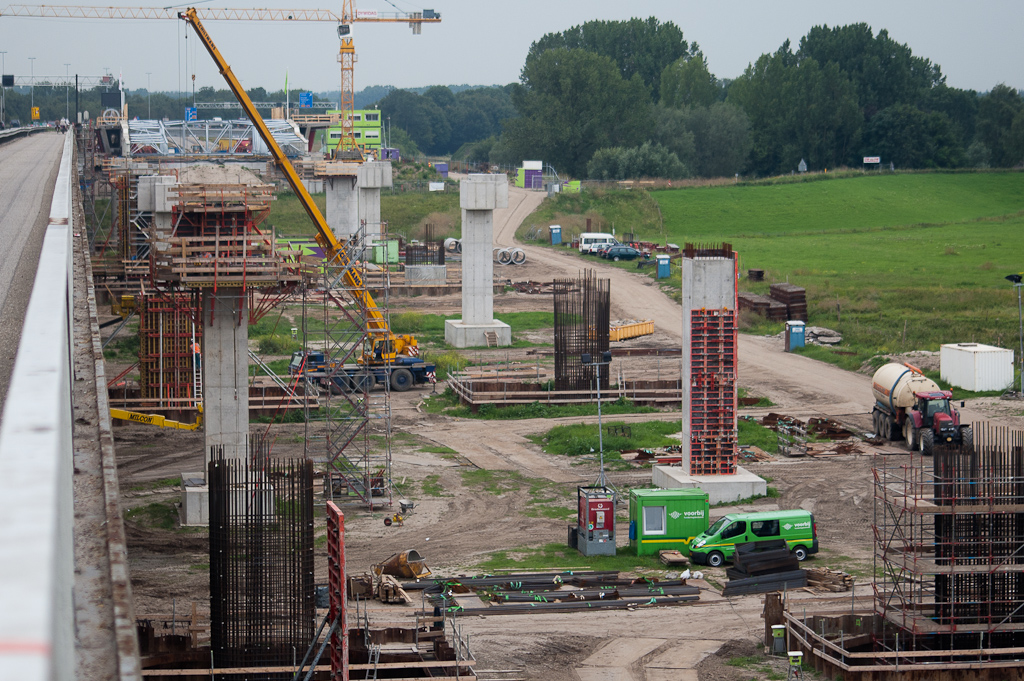 20110803-134223.jpg - Overzicht zuidoever met grootschalig betonconstructiewerk. De voltooide rechthoekige steunen zijn tijdelijk en dienen voor het geleiden van de schuivende brugdekconstructie naar de rivier toe. De ovale exemplaren, waarvan op de voorgrond nog enkel het wapeningsskelet is gerealiseerd, zijn de definitieve brugsteunen.